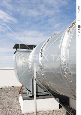 Large Industrial Ventilation System Featuring a Silver Duct with Insulation and a Control Unit, Set Against a Bright Blue Sky with Clouds Large Industrial Ventilation System Featuring a Silver Duct with Insulation and a Control Unit, Set Against a Bright Blue Sky with Clouds 129029661