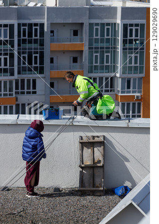 Construction Worker Performing Maintenance on Roof While Child Observes from Below with Curious Eyes Amidst a Modern Urban Setting Construction Worker Performing Maintenance on Roof While Child Observes from Below with Curious Eyes Amidst a Modern Urban Setting 129029690