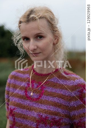 A Portrait of Elegance: A Young Woman with Beautiful Curly Hair Wearing a Stylish Striped Sweater in Earthy Tones Against a Softly Blurred Natural Background 129029968