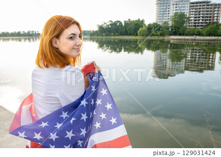 Young womanl holding USA national flag on her shoulders with high office buildings in a city celebrating United States independence day. Young womanl holding USA national flag on her shoulders with high office buildings in a city celebrating United States independence day. 129031424
