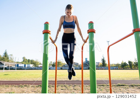 Young sportive athlete woman doing fitness exercise on metal bars outdoors at stadium court. 129031452