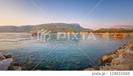 Morning light bathes the Moonlight beach in Kemer, Turkey. Boats rest in the calm bay with turquoise Mediterranean sea water. Lush green hills with palm trees seen at horizon 129031688