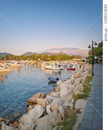 Morning light bathes the Moonlight beach in Kemer, Turkey. Boats rest in the calm bay with turquoise Mediterranean sea water. Lush green hills with palm trees seen at horizon 129031689