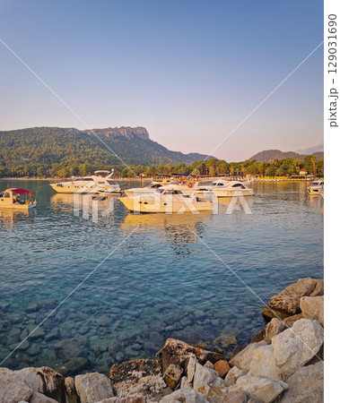Morning light bathes the Moonlight beach in Kemer, Turkey. Boats rest in the calm bay with turquoise Mediterranean sea water. Lush green hills with palm trees seen at horizon 129031690