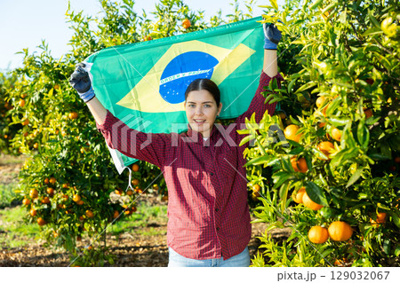 Happy young female gardener waving flag of Brazil during visit tangerine orchard or farm on sunny day Happy young female gardener waving flag of Brazil during visit tangerine orchard or farm on sunny day 129032067