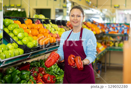Portrait of busy European female seller in apron arranging bell peppers on shelves in grocery supermarket 129032280