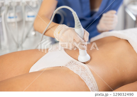 Young female cosmetologist performs ultrasound massage on belly of young woman 129032490