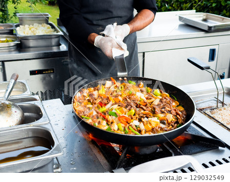 Male chef cooking stir fry outdoors with colorful vegetables and meat in a pan Male chef cooking stir fry outdoors with colorful vegetables and meat in a pan 129032549