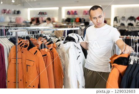 Guy stopped near showcase with outerwear, holds jacket hanger in hands and examines product 129032603