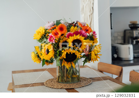 Stylish modern interior of open space white kitchen with huge multicolor summer flower bouquet in vase on wooden kitchen counter table, macrame on the wall. Cozy Design home decor. Selective focus. 129032831