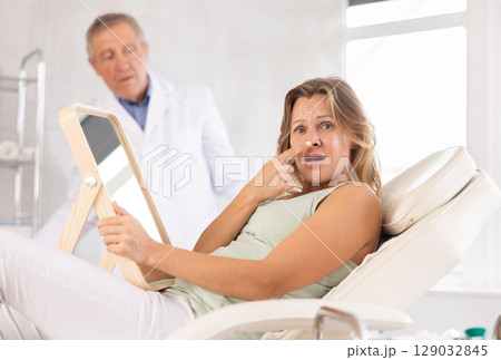 In cosmetology clinic, displeasure client frowns and touches nose, confused doctor in background 129032845