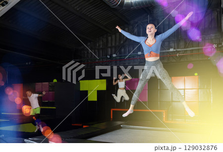 Positive middle aged woman in sporty clothes jumping vertical high in modern trampoline hall 129032876