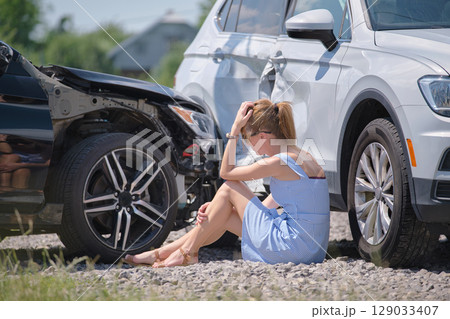 Sad young woman driver sitting near her smashed car looking shocked on crashed vehicles in road accident Sad young woman driver sitting near her smashed car looking shocked on crashed vehicles in road accident 129033407