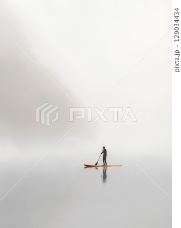 Man paddleboarding on calm water in dense fog 129034434