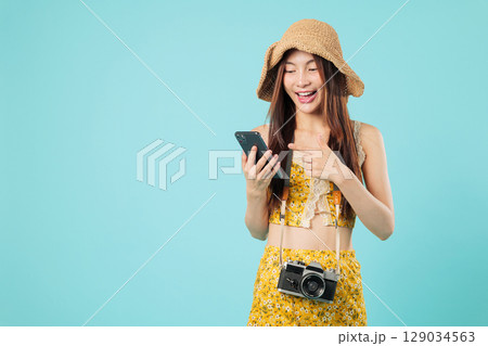 Happy young asian woman travel on summer vacation, a tourist with phone and camera, in clothes and hat is pointing at her mobile screen as passenger tourist, headshots isolated studio blue background 129034563