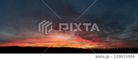 Stunning sunset paints sky with vibrant hues of red, orange, and pink, blending into deep blues and purples. Silhouetted hills create contrast against fiery horizon, adding drama to serene landscape. 129035004