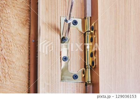 Carpenter installing complete door hinge onto wooden door edge, securing metal hinge with screws, detailed view of installation process shown. 129036153