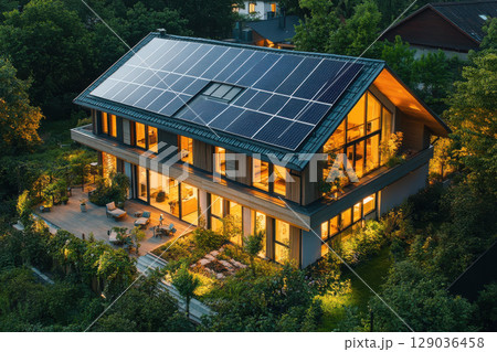 Modern eco-friendly house with solar panels surrounded by lush greenery and evening lights Modern eco-friendly house with solar panels surrounded by lush greenery and evening lights 129036458