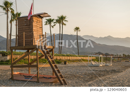 Lifeguard hut on a Greek beach, rescue boat underneath, palm trees and beautiful landscape of Crete Lifeguard hut on a Greek beach, rescue boat underneath, palm trees and beautiful landscape of Crete 129040603