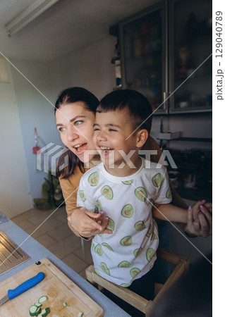 Happy mother hugging her son in the kitchen as they laugh together while preparing fresh cucumber slices. Joyful family bonding during a cooking moment at home. Happy mother hugging her son in the kitchen as they laugh together while preparing fresh cucumber slices. Joyful family bonding during a cooking moment at home. 129040789
