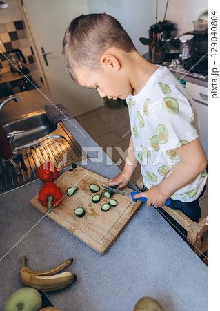 Mother and young boy happily preparing vegetables together in the kitchen. The child is standing on a stool, learning and helping with cucumber slicing on a wooden board Mother and young boy happily preparing vegetables together in the kitchen. The child is standing on a stool, learning and helping with cucumber slicing on a wooden board 129040804