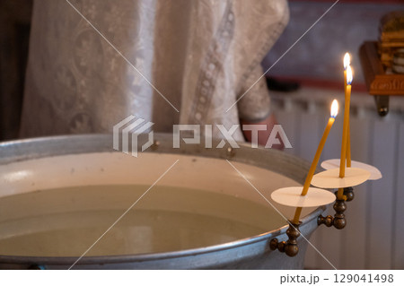 Close-up of a baptismal font filled with holy water and lit ceremonial candles, used during an Orthodox Christian baptism ritual 129041498