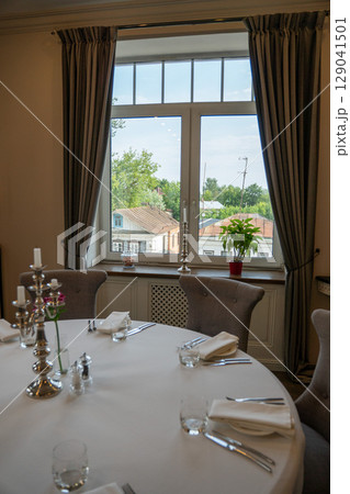 Elegant restaurant interior with a round table set for six near a large window overlooking old town rooftops and trees Elegant restaurant interior with a round table set for six near a large window overlooking old town rooftops and trees 129041501
