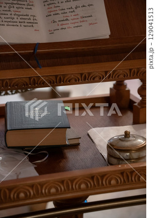 Close-up of an Orthodox prayer book, placed on a carved wooden lectern inside a church, accompanied by a small ornate container and baptismal crosses 129041513