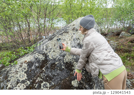 A woman photographs a moss-covered boulder in a lush northern forest using her smartphone 129041543