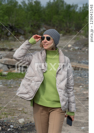 Woman in a light down jacket, green sweater, and grey beanie walks outdoors on a cloudy day, smiling and adjusting her sunglasses 129041548