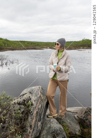 Woman in a beige puffer jacket and gray beanie stands by an Arctic lake holding a turquoise takeaway coffee cup 129041565