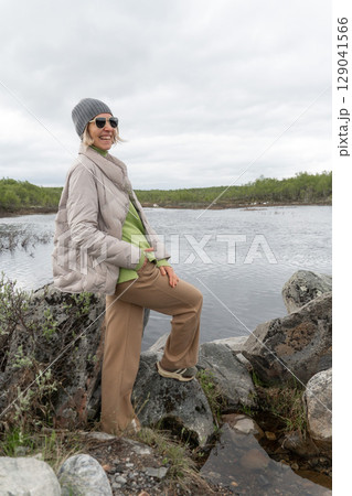 Woman in casual outdoor clothing smiles joyfully while standing on rocky terrain beside a cold northern lake in Teriberka, Russia 129041566