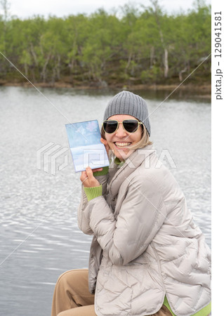 Smiling woman in warm clothes poses by a northern lake in Teriberka, holding a polar explorer passport in her hand 129041581