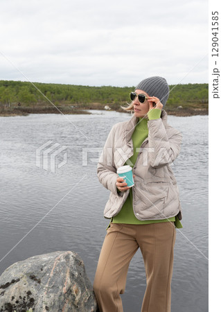 Woman in a beige puffer jacket and gray beanie stands by an Arctic lake holding a turquoise takeaway coffee cup Woman in a beige puffer jacket and gray beanie stands by an Arctic lake holding a turquoise takeaway coffee cup 129041585