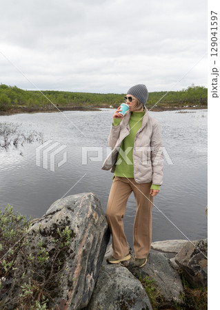 Woman in beige jacket and gray beanie drinks from a turquoise coffee cup while standing on rocks by a northern lake in Teriberka, Russia 129041597