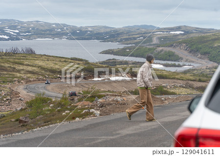 A woman in a gray beanie and beige jacket crosses a winding road surrounded by the Arctic tundra landscape near Teriberka, Russia A woman in a gray beanie and beige jacket crosses a winding road surrounded by the Arctic tundra landscape near Teriberka, Russia 129041611