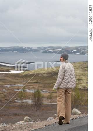 Woman in beige jacket and gray beanie walking along a road overlooking Arctic tundra and Barents Sea with snow patches on distant hills 129041627