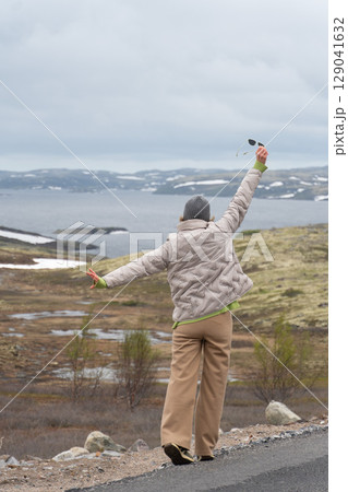Joyful woman in beige jacket and gray beanie raising arms with sunglasses in hand, enjoying the dramatic Arctic scenery with snowy hills 129041632