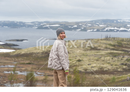 Smiling woman in beige jacket and gray beanie standing on Arctic tundra with Barents Sea and snow-covered hills in background Smiling woman in beige jacket and gray beanie standing on Arctic tundra with Barents Sea and snow-covered hills in background 129041636