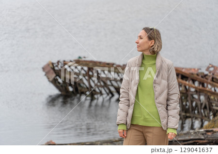 Woman in beige jacket and green sweater standing near a rusted shipwreck in calm northern waters, looking thoughtfully into the distance 129041637