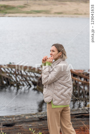 Woman in a beige puffer jacket and sunglasses standing near rusted shipwrecks by the shore, looking at the camera with a calm expression 129041638