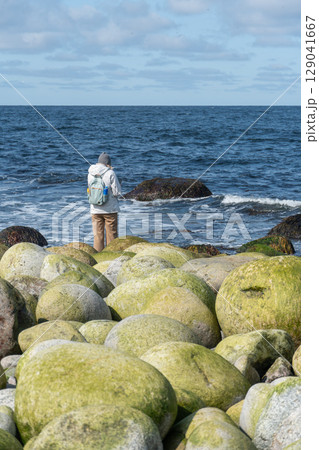 Woman in a white jacket standing on Dragon Eggs Beach, Teriberka, facing the Barents Sea with green algae-covered round boulders in the foreground 129041667