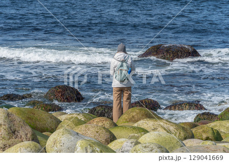 Woman in a gray beanie and white jacket with a backpack stands on the rocky Dragon Egg Beach, facing the Barents Sea waves 129041669