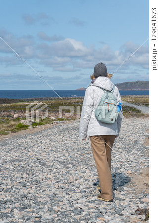 Woman in a white jacket and gray beanie walking along a rocky coastal path in Teriberka nature reserve with the Barents Sea and cliffs Woman in a white jacket and gray beanie walking along a rocky coastal path in Teriberka nature reserve with the Barents Sea and cliffs 129041673