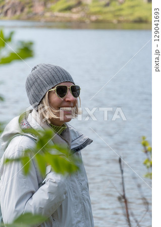 Smiling woman in sunglasses and gray beanie stands by a calm lake, dressed in light outdoor clothing with a backpack, framed by soft green foliage Smiling woman in sunglasses and gray beanie stands by a calm lake, dressed in light outdoor clothing with a backpack, framed by soft green foliage 129041693