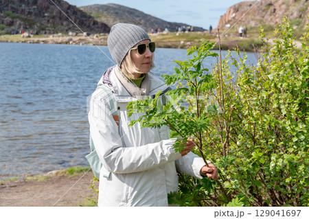 Woman in sunglasses and gray beanie stands by a green bush near a mountain lake, dressed in light outdoor clothing with a backpack Woman in sunglasses and gray beanie stands by a green bush near a mountain lake, dressed in light outdoor clothing with a backpack 129041697