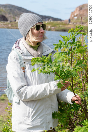 Woman in sunglasses and gray beanie stands by a green bush near a mountain lake, dressed in light outdoor clothing with a backpack 129041706