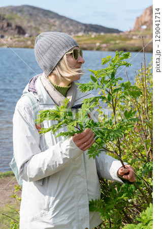 Woman in sunglasses and gray beanie stands by a green bush near a mountain lake, dressed in light outdoor clothing with a backpack Woman in sunglasses and gray beanie stands by a green bush near a mountain lake, dressed in light outdoor clothing with a backpack 129041714