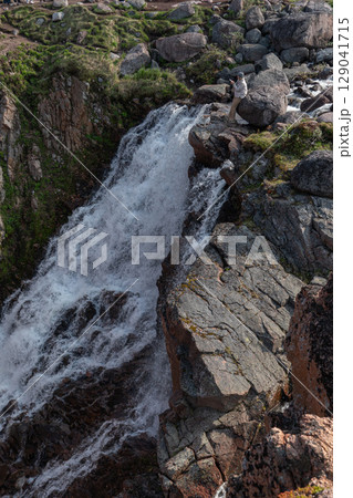 Woman standing on rocks near the powerful Battery waterfall, capturing the scene with her smartphone against rugged Arctic nature 129041715