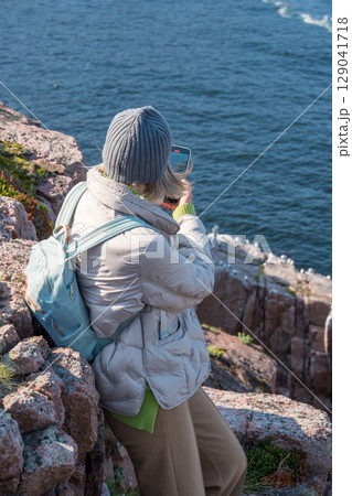 Woman in warm clothing sitting on a rocky Arctic cliff at the Barents Sea in Teriberka park, photographing a seabird colony with her smartphone 129041718